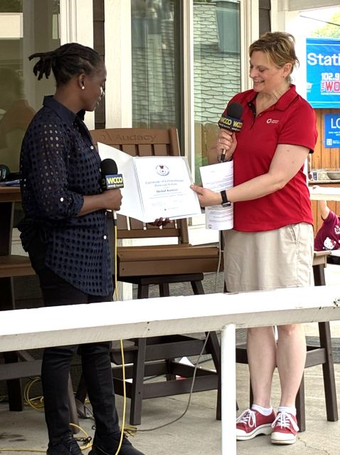 Two women speaking at an outdoor event, one holding a certificate and the other holding a microphone.