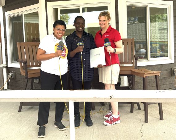 Three individuals posing together outdoors, each holding a microphone. The woman on the left is wearing a white shirt, the man in the middle is dressed in a black shirt with patterns, and the woman on the right is in a red shirt, holding a clipboard. They are in front of two wooden chairs and a window.